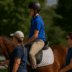 Two instructors walk beside a mounted rider during an adaptive riding session, guiding a calm bay horse in a sunny outdoor arena surrounded by green trees.