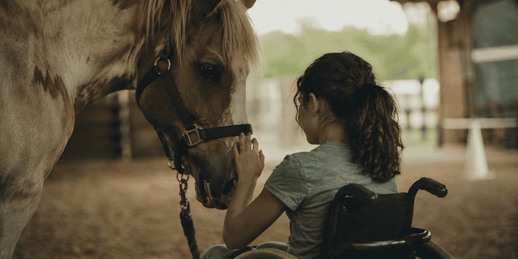 Child in a wheelchair gently touching a therapy horse in a quiet indoor riding arena.