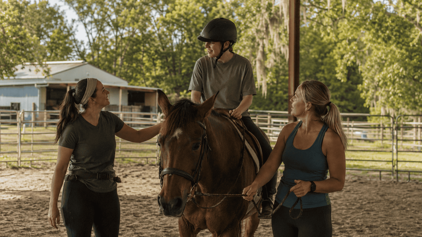 A natural adaptive riding lesson with instructors supporting a mounted rider in a covered outdoor arena.